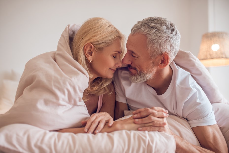 A imagem mostra um casal de meia-idade em um momento íntimo e carinhoso, deitados na cama e trocando olhares afetuosos sob um cobertor. Esse registro ilustra que, mesmo com o avanço da idade, é possível manter uma vida sexual ativa e satisfatória. Muitas pessoas se perguntam se quem tem problema de próstata pode ter relação, e a resposta é sim.