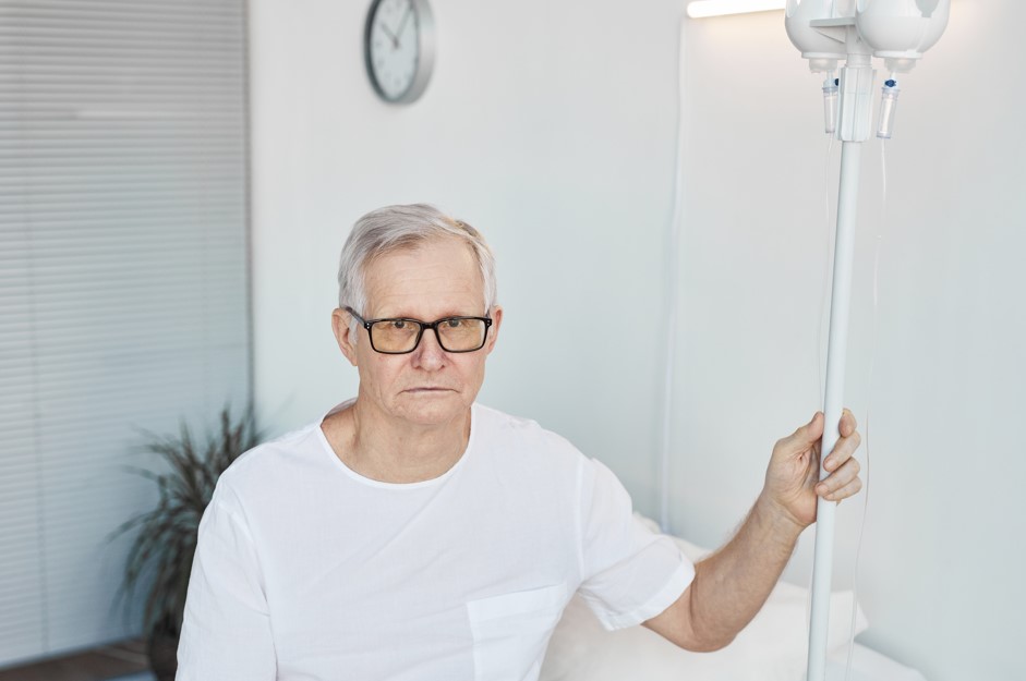 Paciente, homem por volta dos 65 anos, sentado em uma cama de hospital após passar por uma ressecção transuretral da próstata.