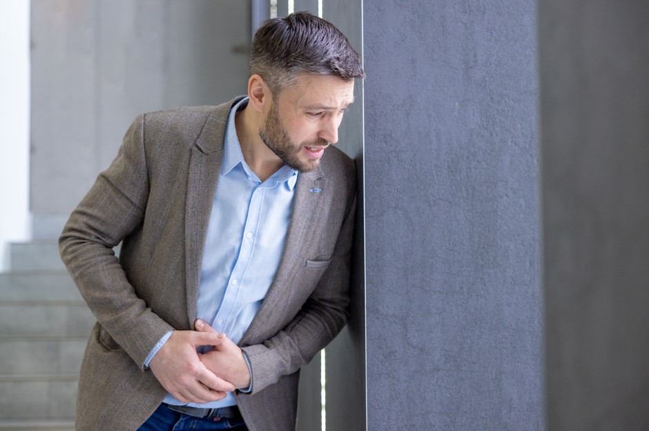 Um homem de meia-idade, vestindo um blazer cinza e uma camisa azul, está visivelmente desconfortável, com uma expressão de dor enquanto segura a parte inferior do abdômen. Esta situação é típica de alguém que pode estar experienciando sintomas de uma infecção urinária, uma condição que geralmente requer a intervenção de especialistas em infecção urinária para tratamento adequado e alívio efetivo dos sintomas.