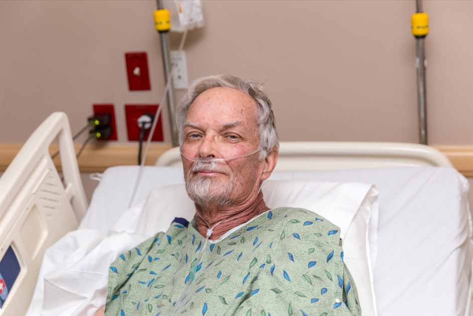 Um homem idoso com barba branca está sorrindo enquanto está sentado em uma cama de hospital, usando uma bata hospitalar estampada e recebendo oxigênio através de um tubo nasal. Esta cena pode ilustrar um paciente em recuperação ou em tratamento contínuo, talvez relacionado a condições como o câncer de próstata.