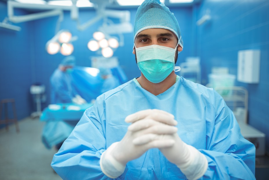 Cirurgião com luvas esterilizadas e uma máscara cirúrgica, enquanto faz um gesto com as mãos unidas, possivelmente se preparando para iniciar uma cirurgia de próstata a laser.