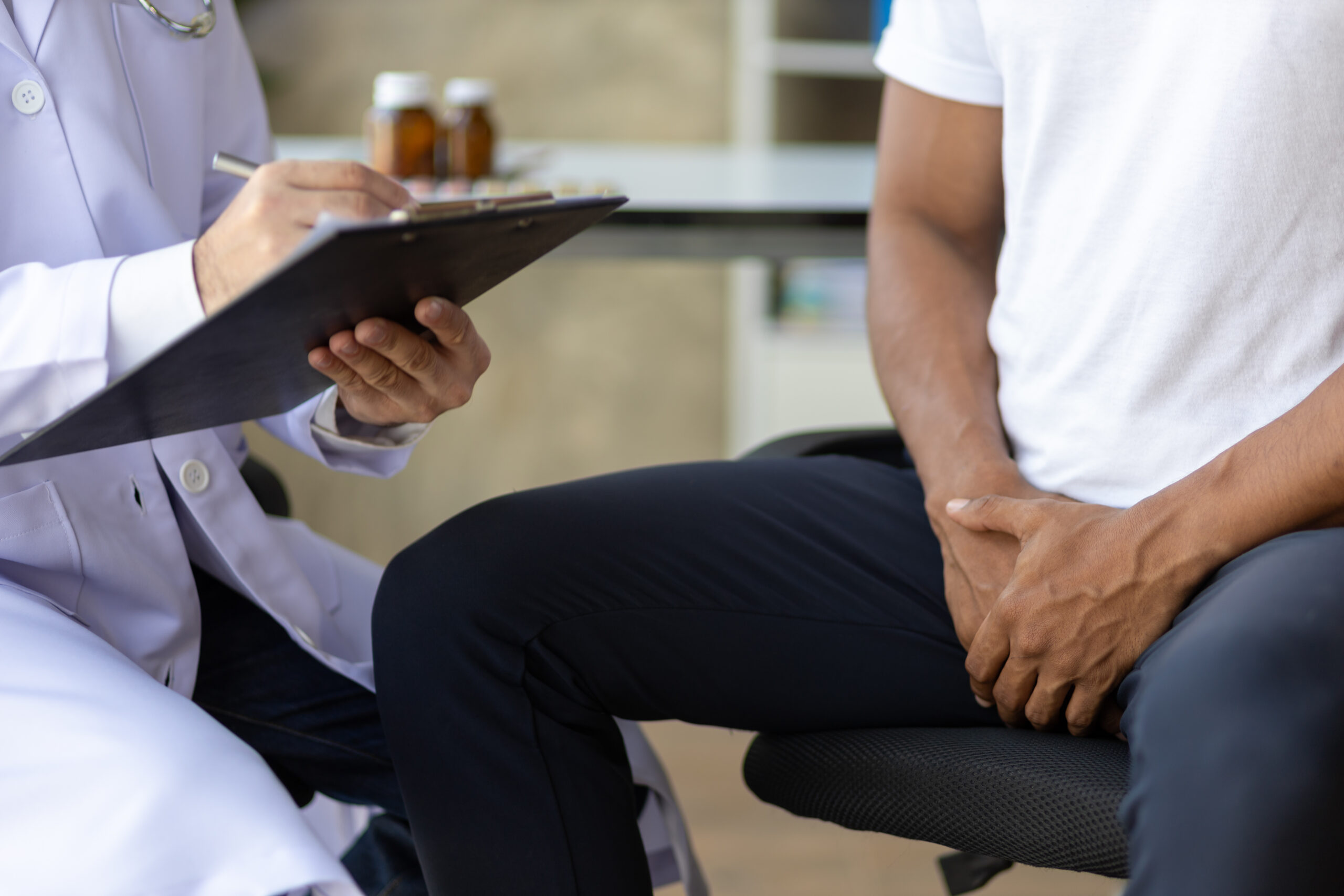 Profissional de saúde anotando informações em prancheta durante consulta, enquanto paciente sentado segura o abdômen, indicando possível dor, em ambiente de consultório médico.