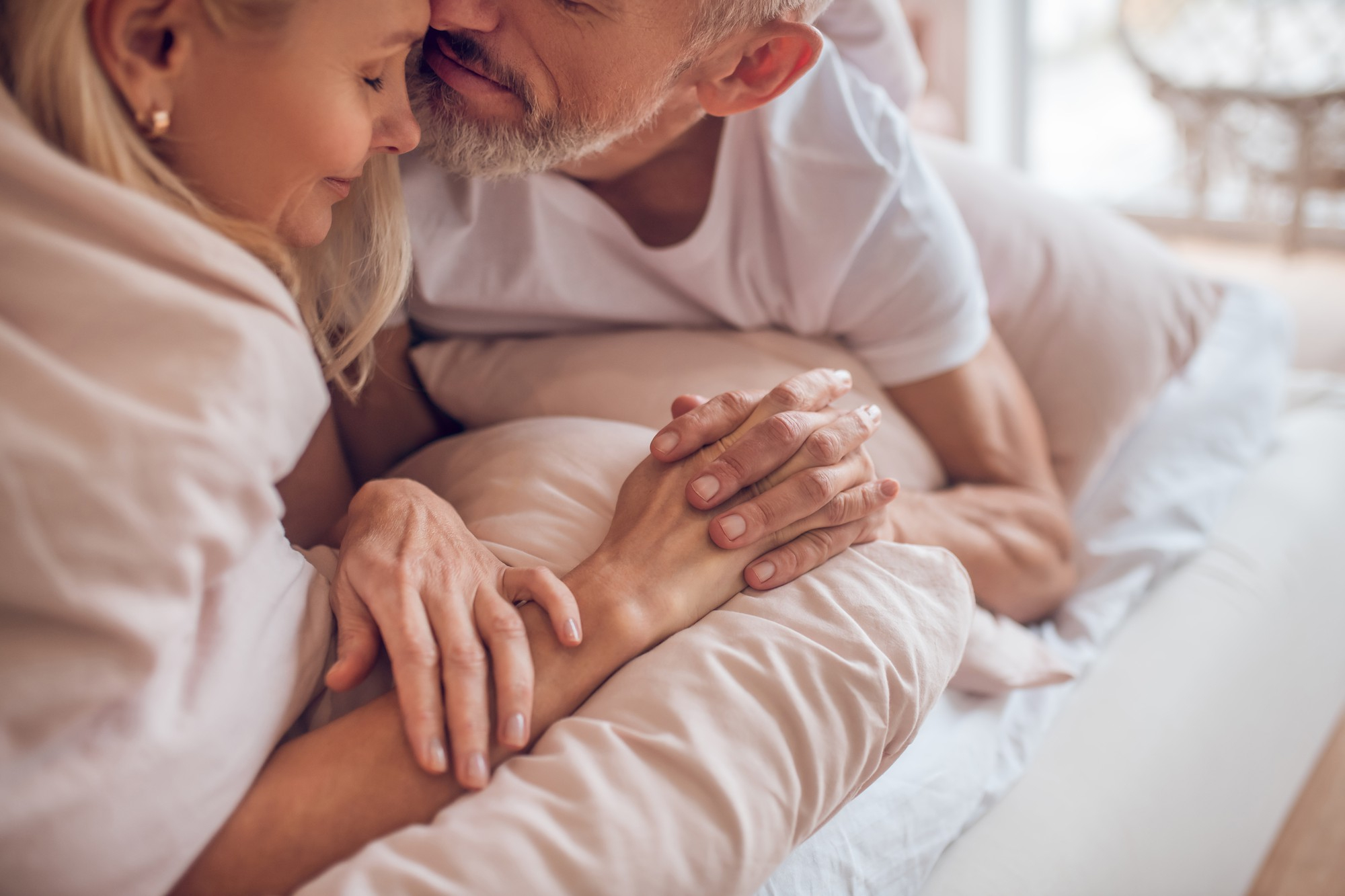 A imagem mostra um momento íntimo de um casal maduro deitado na cama, onde o homem se inclina para a mulher em um gesto de carinho relacionado ao tema “quem tem câncer de próstata pode ter relações?”. O momento é de ternura e conforto, enfatizado pelas mãos entrelaçadas no centro, transmitindo a conexão e a intimidade do casal.