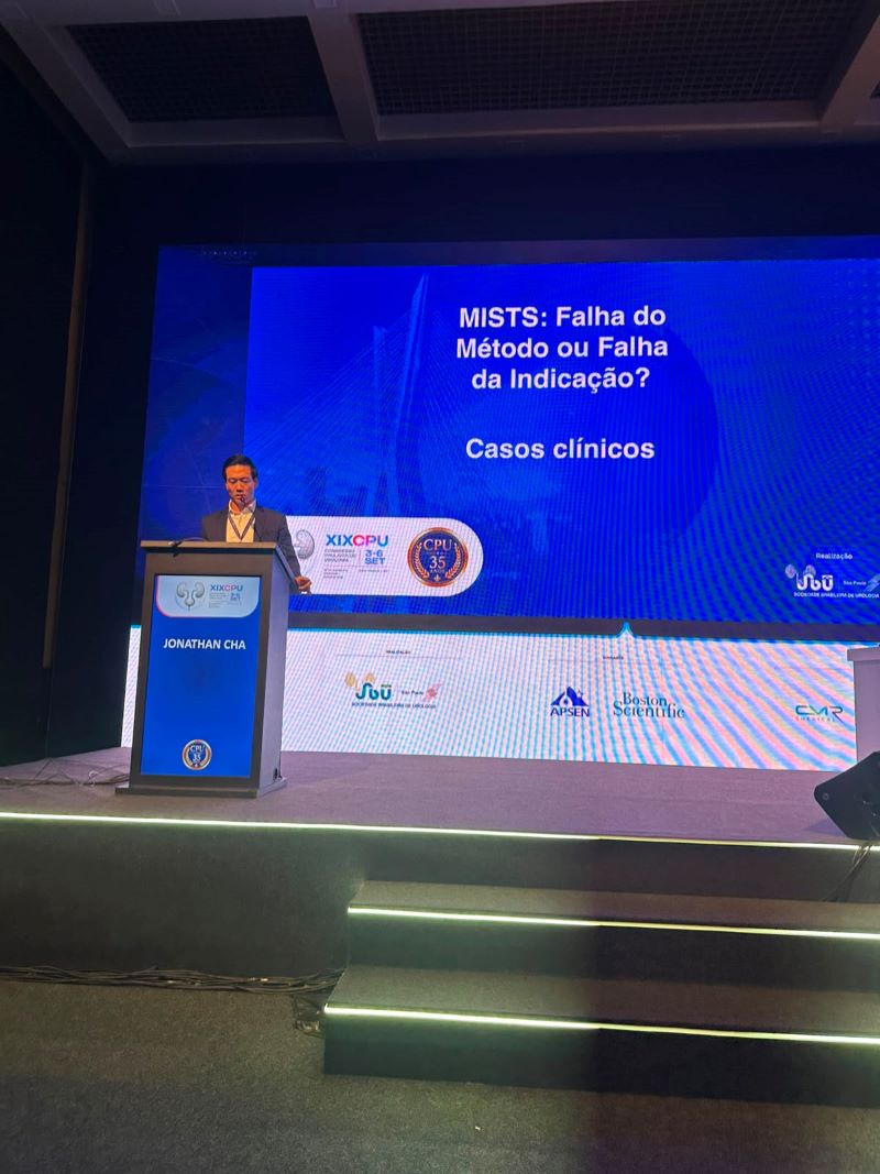 Dr. Jonathan Cha apresenta casos clínicos durante o Congresso Paulista de Urologia 2026, em um palco com telão ao fundo.